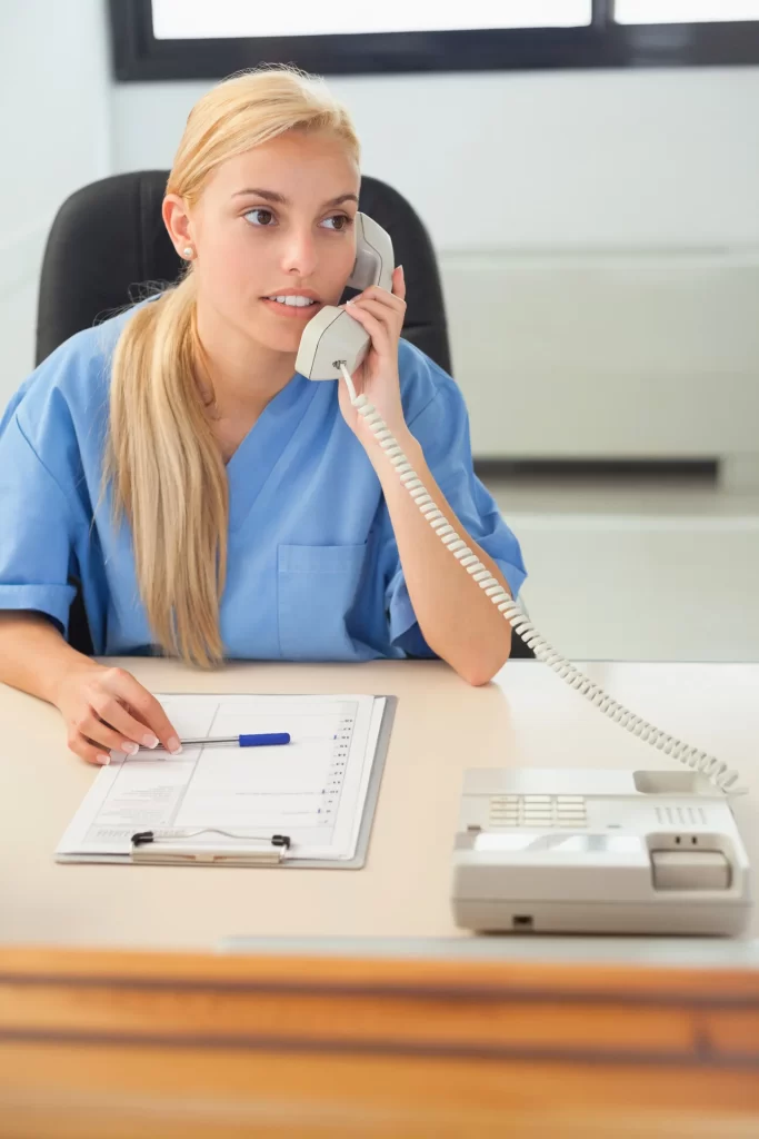 Žena v modrých peelingech sedí u psacího stolu v Ústí nad Labem a mluví do provázaného telefonu. Nad schránkou drží propisku, která ztělesňuje klidné zaměření zdravotní péče, která se často vyskytuje v ordinaci praktického lékaře.